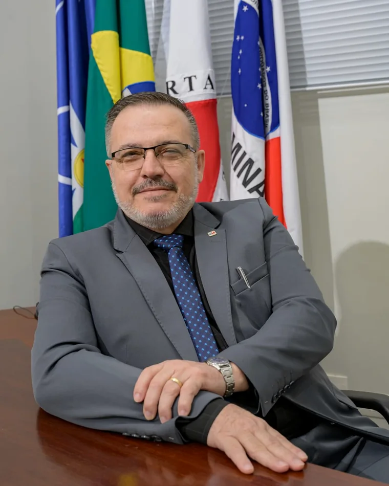 Dr. Vilmar Cunha sentado à mesa de reuniões, diante das bandeiras do Brasil e de Minas Gerais